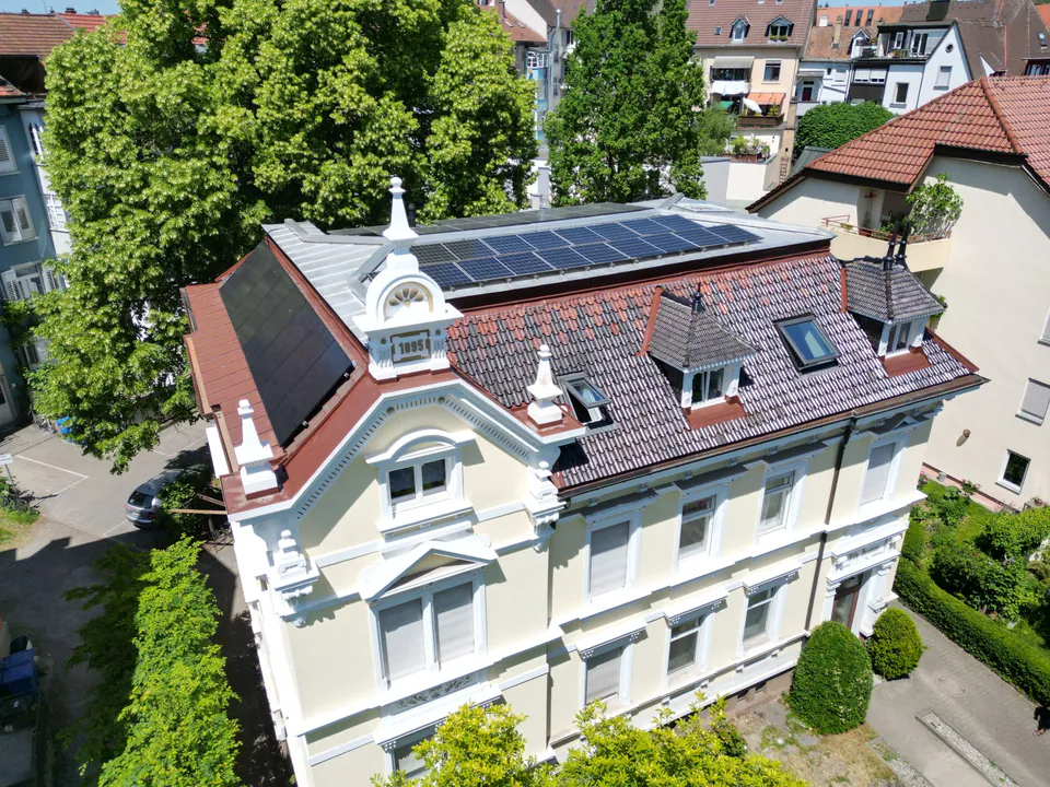 Altbau mit heller Fassade und verziertem Giebel, von schräg oben gesehen: schwarze Solarmodule in der steilen Dachfläche, weitere Module auf dem flachen Dachteil.