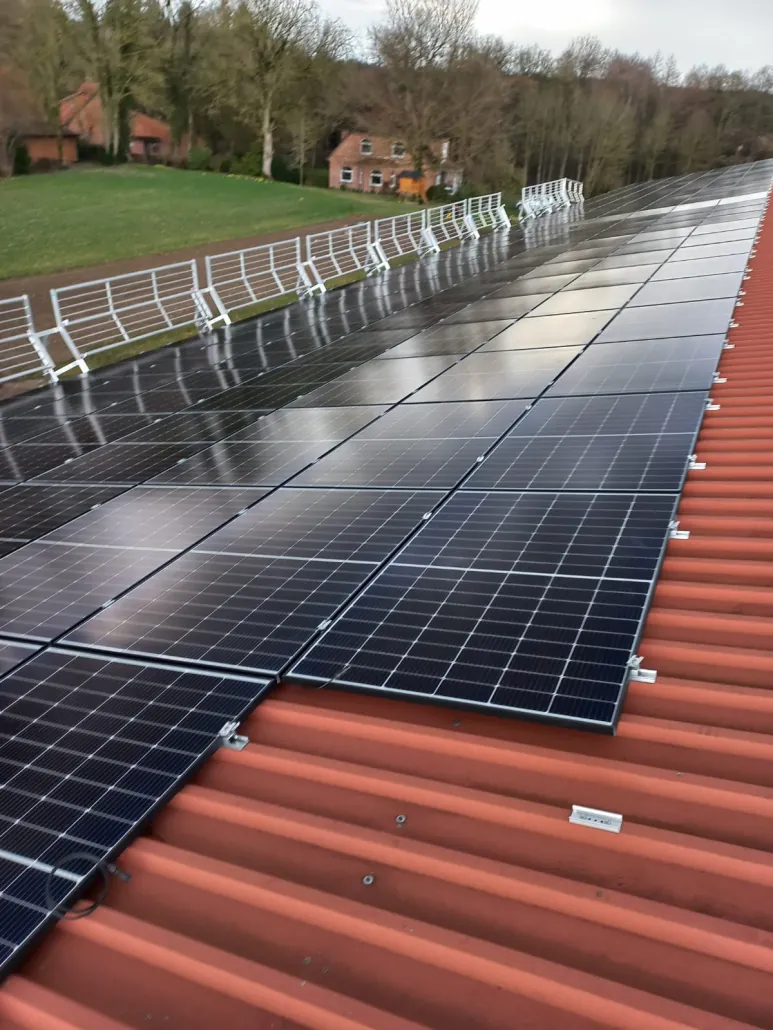 Nahaufnahme: Solarmodule in Reihen auf einem roten Blechdach, am Dachrand ein weisses Geländer, dahinter Wiese, Bäume und Backsteinhäuser.