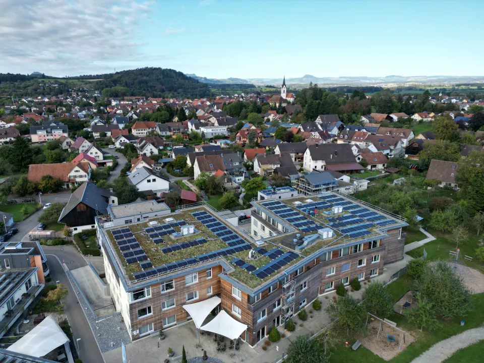 Luftaufnahme: mehrstöckiges Gebäude mit Holzfassade und begrüntem Flachdach mit Solarmodulen, dahinter ein Dorf mit Kirchturm und Hügel am Horizont.