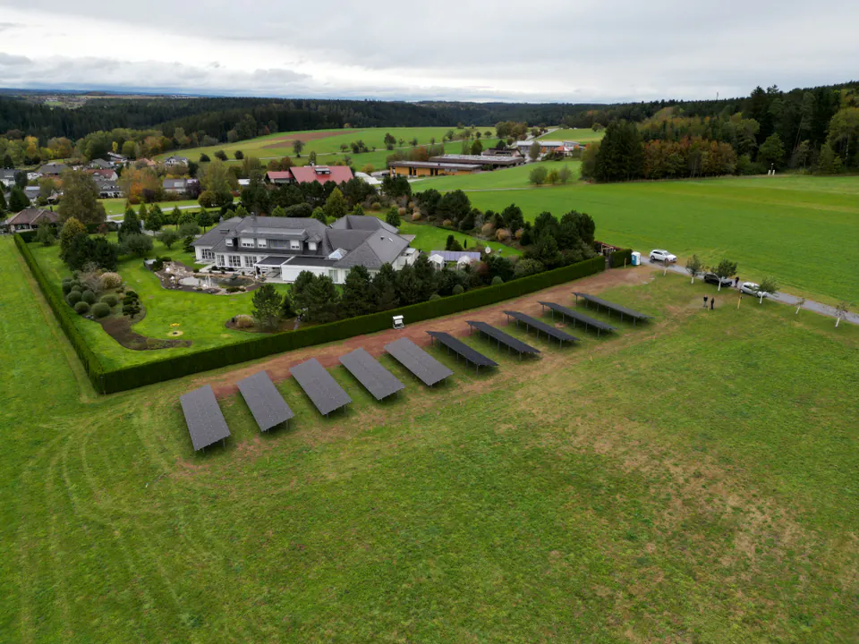 Luftaufnahme: eine Reihe aufgeständerter Solarmodule auf der Wiese neben einem Wohnhaus mit Garten und Hecke, im Hintergrund Felder, Dorf und Wald.